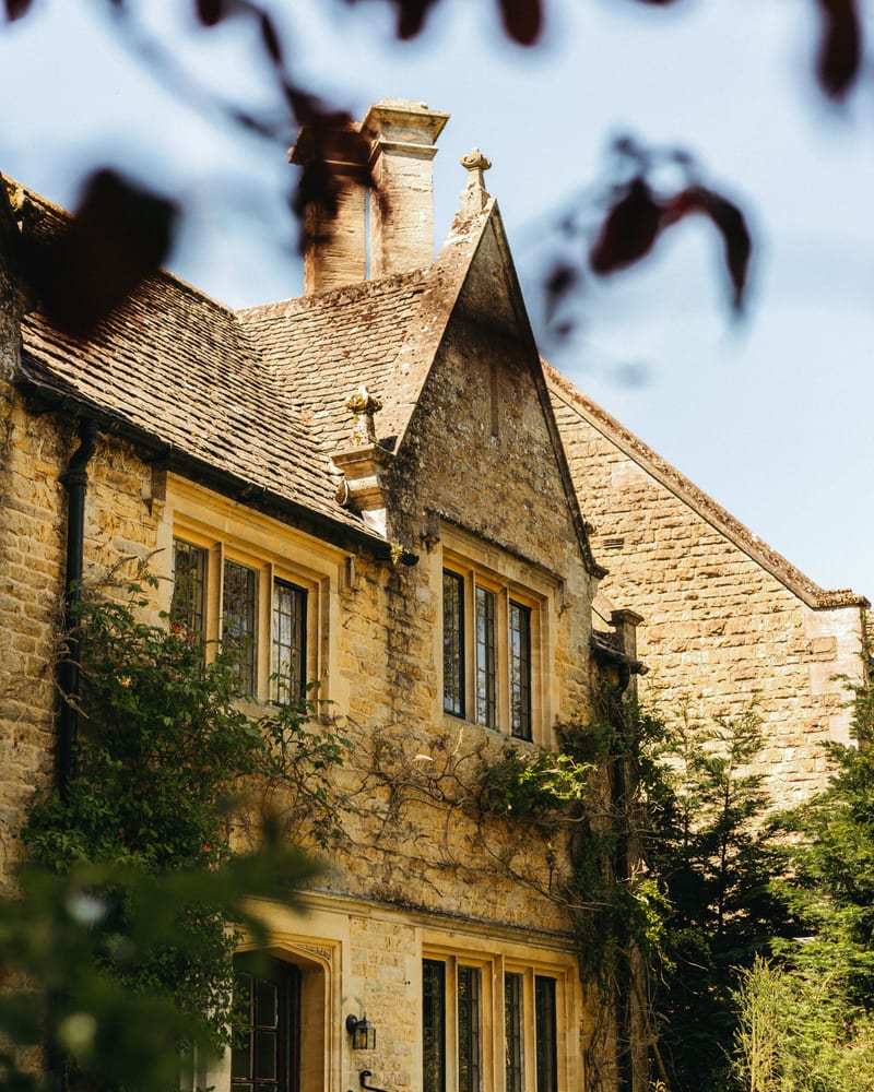 A pale stone building with mullioned windows, chimneys and climbing plants, framed by trees.